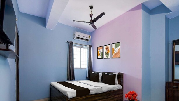 Un dormitorio con cama, ventilador de techo, unidad de aire acondicionado, ventana con cortinas y una mesita con un jarrón de flores.