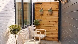 Un petit patio extérieur avec un sol en pierre, deux chaises blanches, une plante en pot et une clôture en bois.