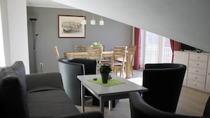 A modern living room with a dining area, a sofa, a table with a plant, and a floor lamp. - Ferienwohnung in Orth auf Fehmarn (Fehmarn)