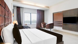 A modern hotel room with a large bed, a flat-screen TV mounted on the wall, a seating area with a chair and table, and a window with curtains.