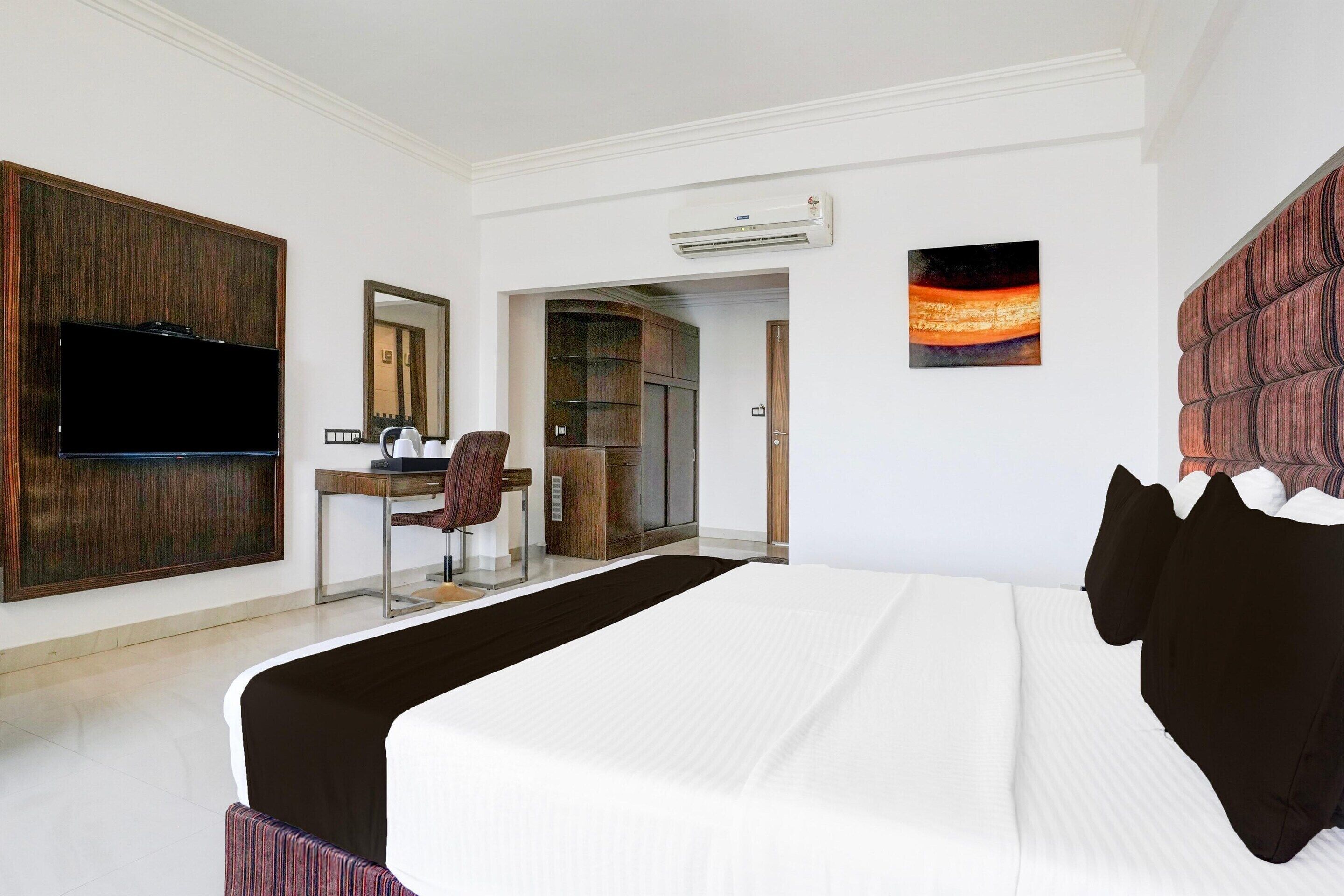 A hotel room with a large bed, a desk with a chair, a television, a wardrobe, and an air conditioning unit.