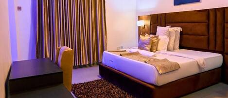 Habitación de hotel con cama, escritorio, silla, ventana con cortinas y una obra de arte colgada en la pared.