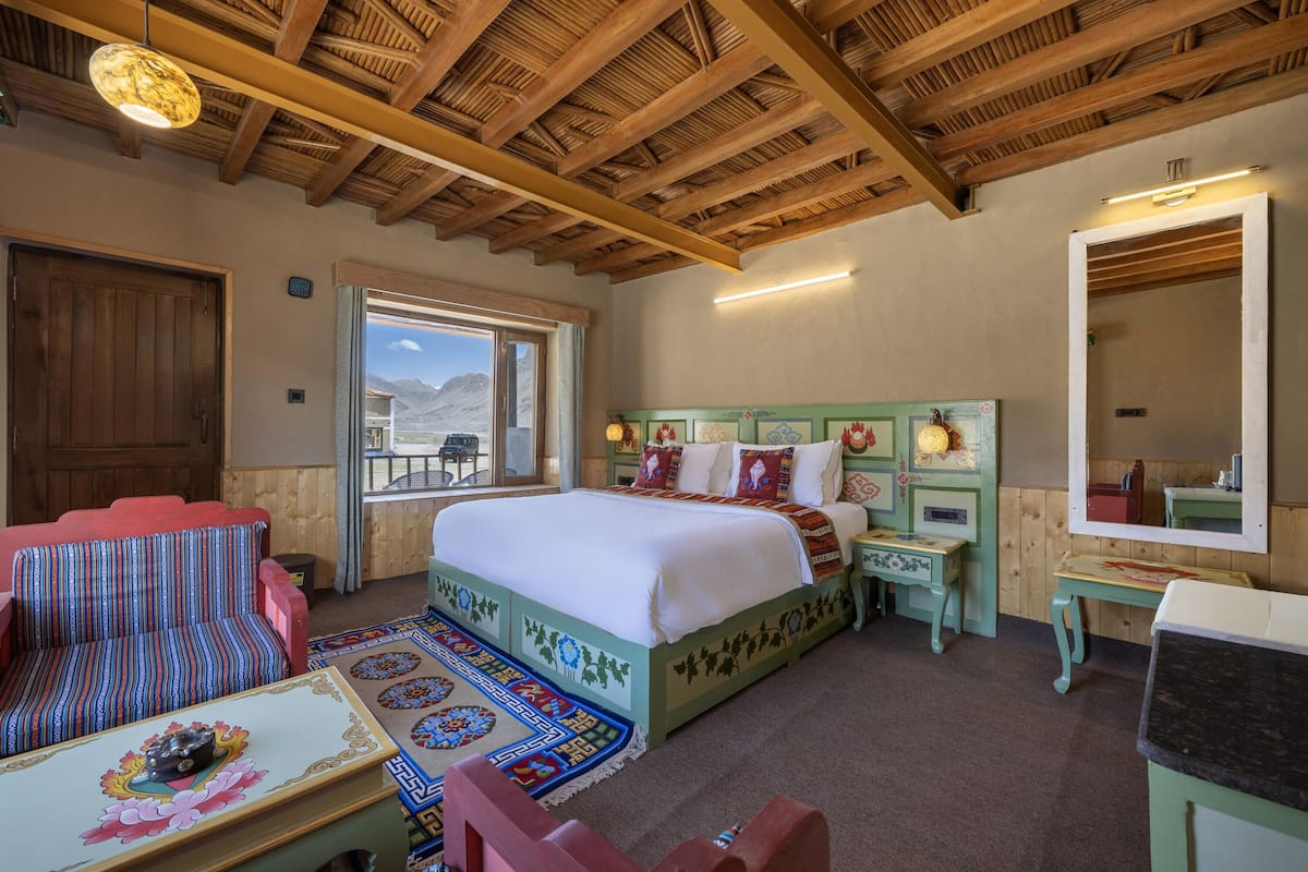 A room with a wooden ceiling, a bed with a colorful headboard, a bench, a small table, and a mirror.