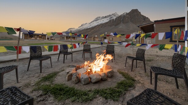 Een vuurkuil met rondom geplaatste stoelen, een gebouw met een berg op de achtergrond, en kleurrijke vlaggen tussen de stoelen gespannen.
