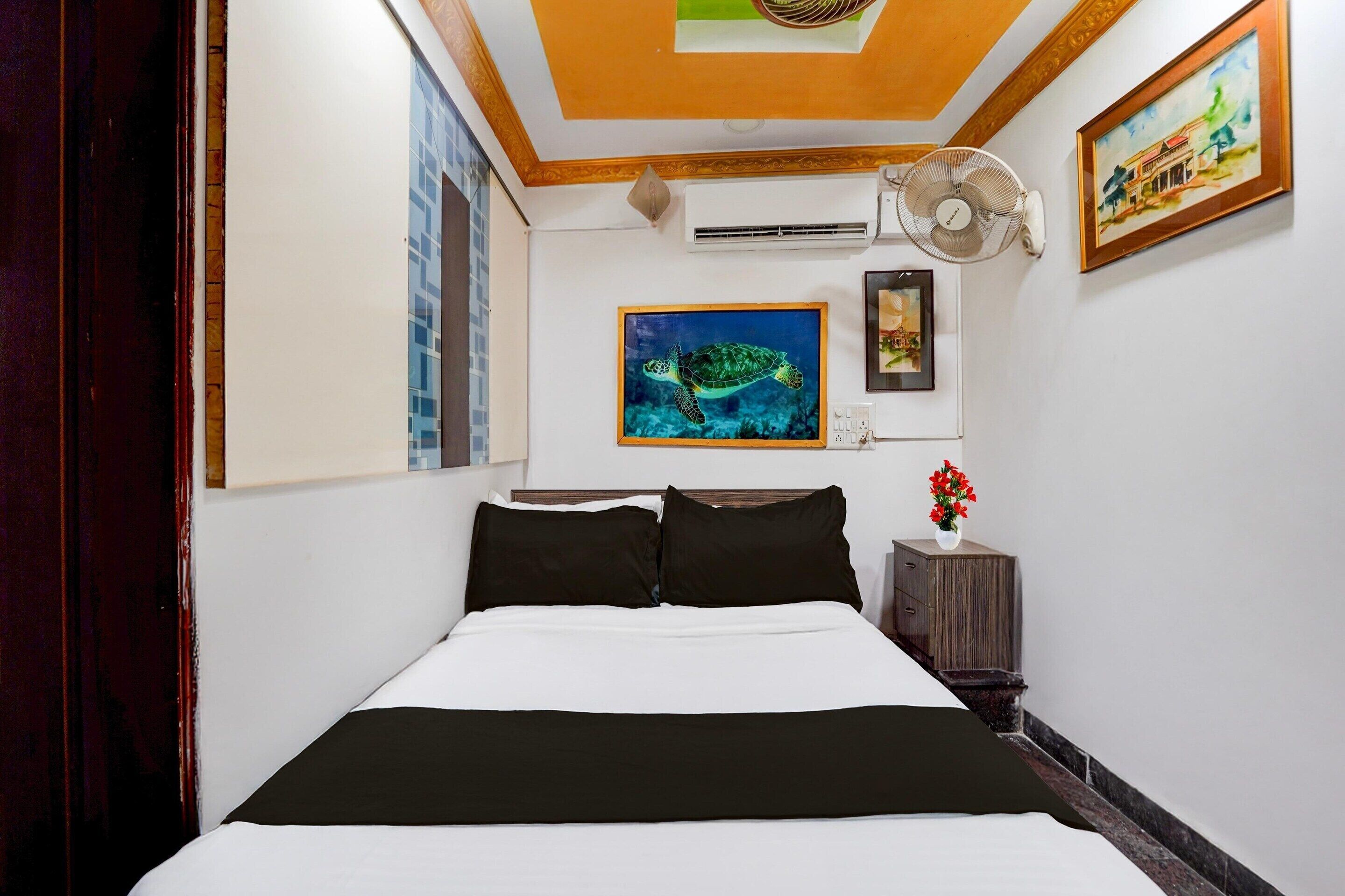 A hotel room with a bed, a wall-mounted air conditioner, a ceiling fan, a painting of a turtle, and a small table with a vase of flowers.