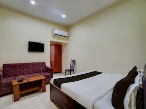A bedroom with a bed, a wooden nightstand, a red sofa, a TV mounted on the wall, and an air conditioning unit. - Deluxesamne Ghat (Varanasi)