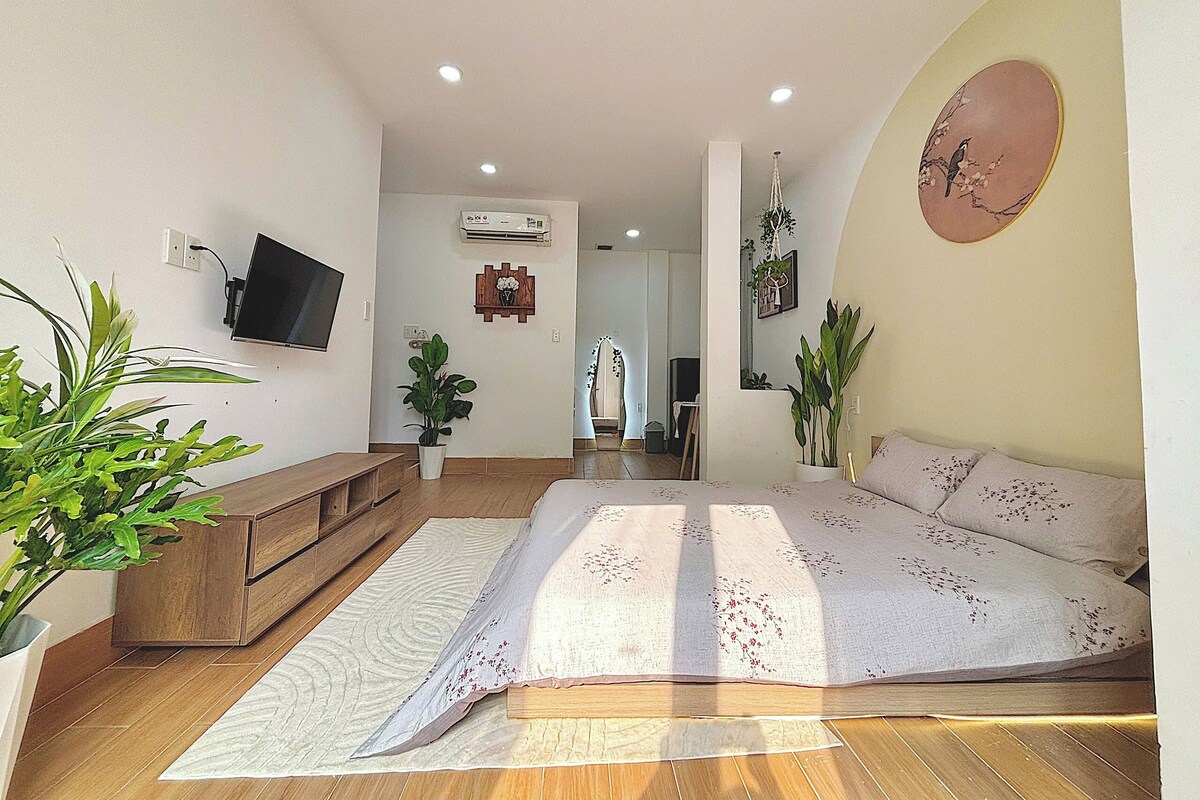 A spacious living room with a large bed, wooden furniture, and several potted plants.