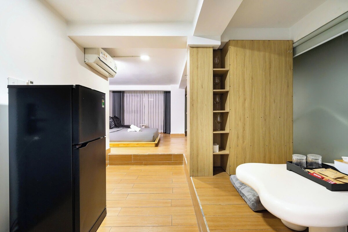 A modern hotel room with a black refrigerator, a bed with a wooden platform, a wooden wardrobe, and a small seating area with a tray of items.