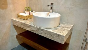 Un baño con un lavamanos blanco redondo, encimera de mármol, tocador de madera y un perchero con una planta.