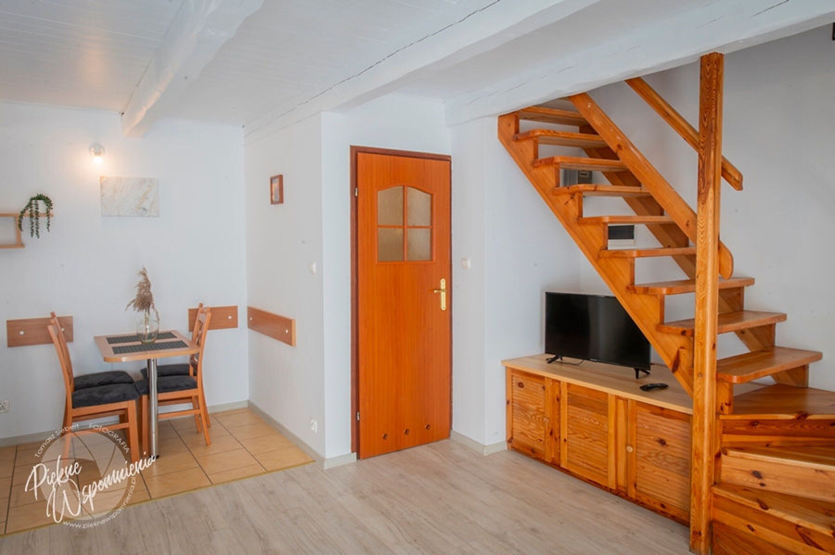 Ein Zimmer mit einer Holztreppe, einer Tür, einem Fernseher auf einem Holzschrank und einem kleinen Tisch mit Stühlen.