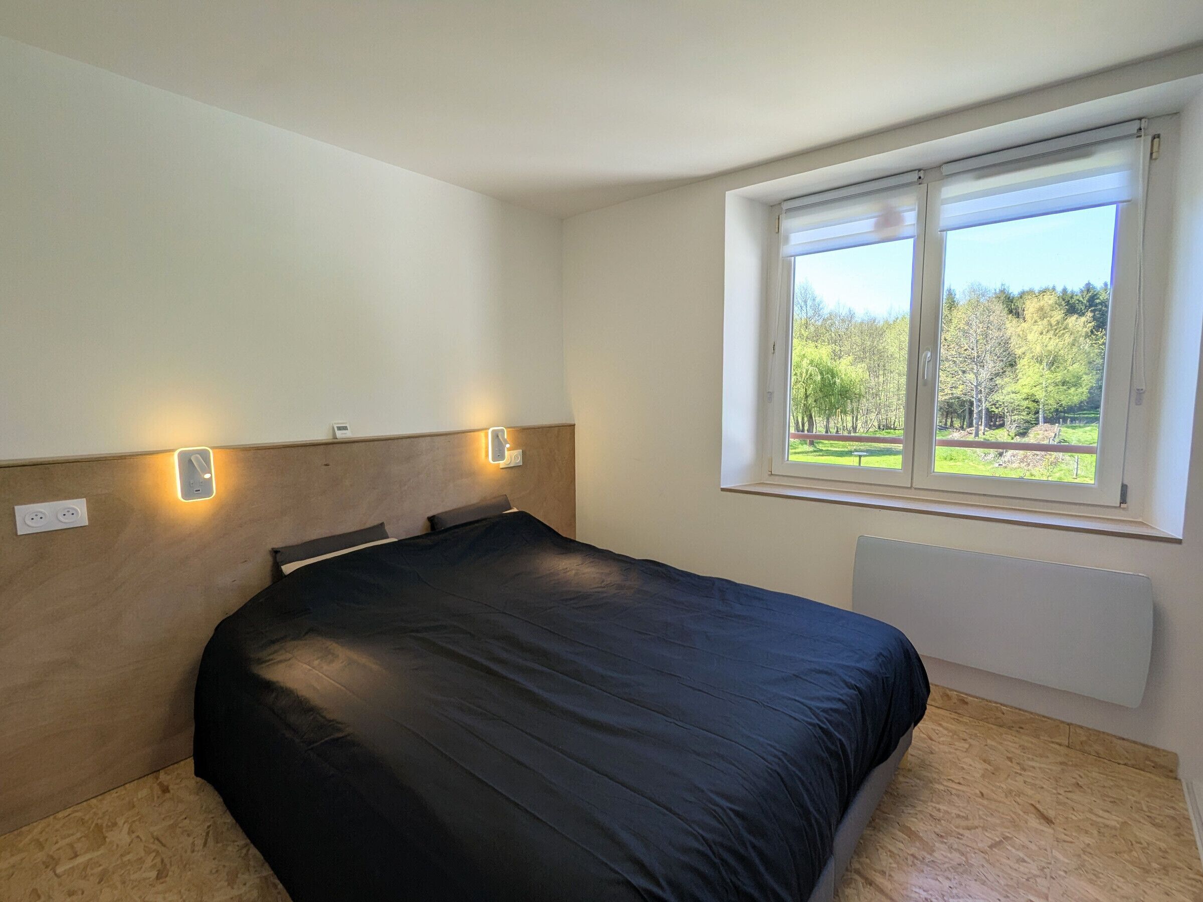 A bedroom with a bed, two bedside lamps, a window with a view of greenery, and a radiator.