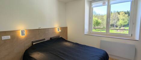 A bedroom with a bed, two bedside lamps, a window with a view of greenery, and a radiator.