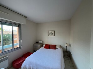 Un dormitorio con cama, ventana, mesita de noche, lámpara y un cuadro en la pared.