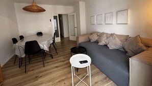 A living room with a sofa, a dining table with chairs, a hanging light fixture, and a door leading to another room. - Renovated house near the beach - 8 guests (Roses)