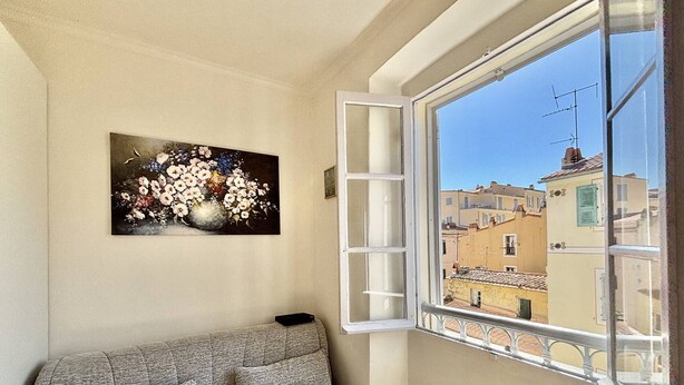 Ein Zimmer mit grauem Sofa, einem runden Holztisch, einem Blumenbild und einem Fenster mit Blick auf Gebäude.