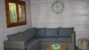 A room with a grey sectional sofa, a small wooden table, a wicker chair, a wall clock, and a window with wooden shutters. - Cozy chalet - 6 guests (Roche-des-Arnauds)