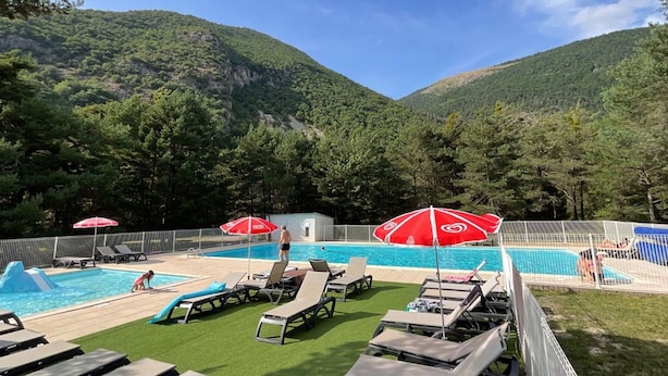 A pool area with lounge chairs and umbrellas, surrounded by a fence and trees, with a mountain in the background. - Cozy chalet - 6 guests (Roche-des-Arnauds)