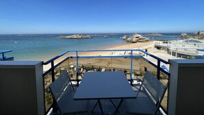 Een balkon met een tafel en stoelen, met uitzicht op een strand met rotsen en boten.