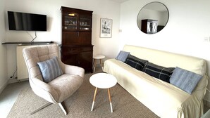 Een woonkamer met een bank, fauteuil, televisie, kast en een ronde spiegel.
