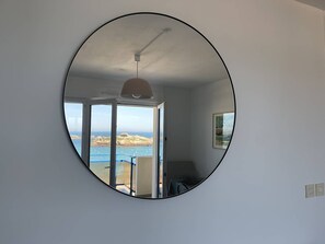 A room with a large circular mirror, a ceiling fan, a door leading to an outdoor area with a view of the sea and rocks, and a small table with a chair. - Apartment exceptional sea view - 3 guests (Trégastel)