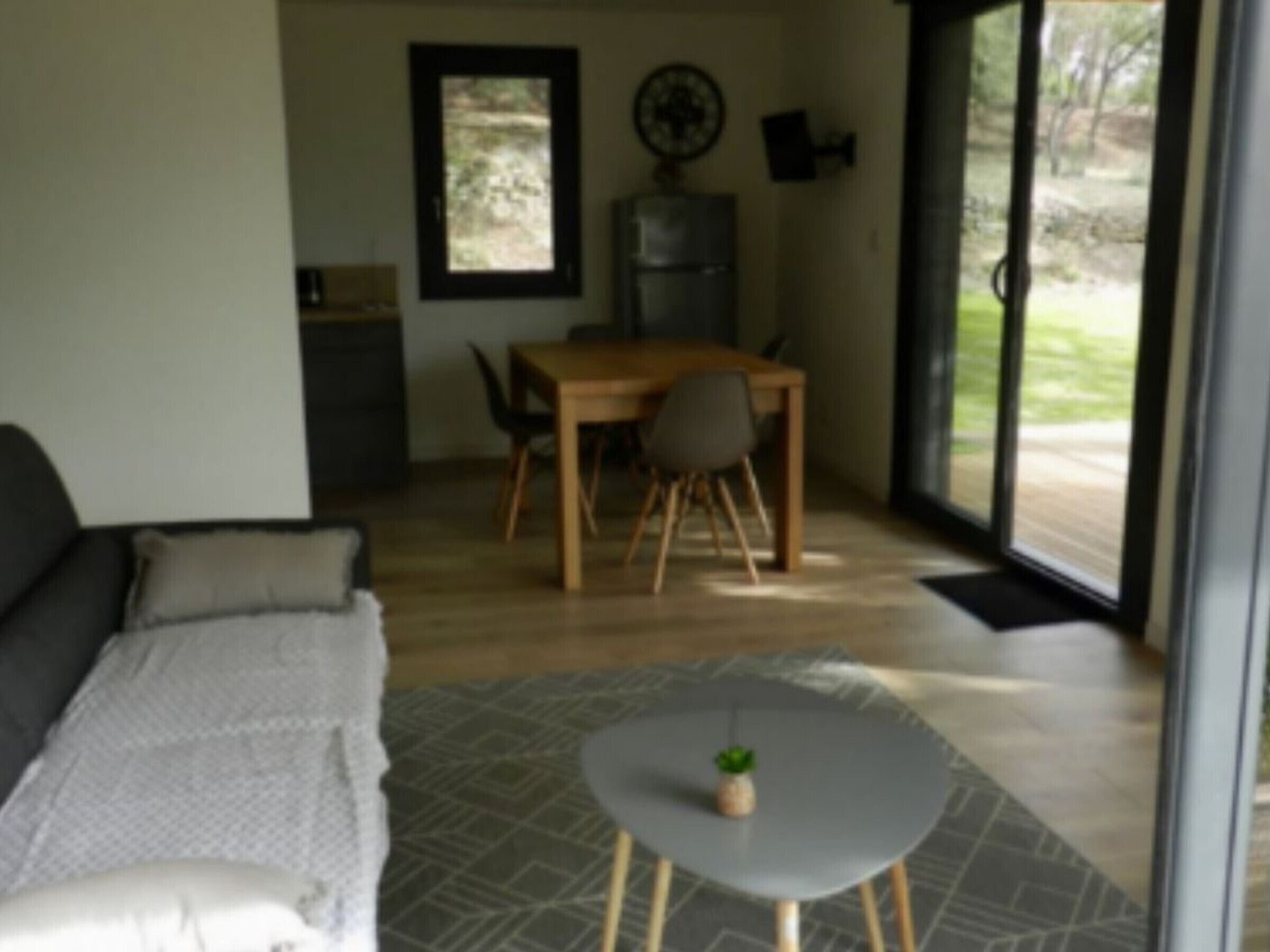 Ein modernes Wohnzimmer mit einer Couch, einem kleinen runden Tisch mit einer Pflanze, einem Essbereich mit Tisch und Stühlen sowie Blick ins Freie durch große Glasfenster.