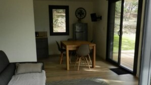 A modern living room with a sofa, a small round table with a plant, a dining area with a table and chairs, and a view of the outdoors through large glass doors. - Chalet with AC - 5 guests - Ventoux camping (Bollène)