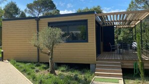 A modern single-story building with a wooden exterior, a small tree in front, and a covered outdoor seating area. - Chalet with AC - 5 guests - Ventoux camping (Bollène)