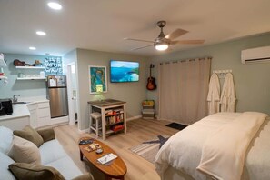 Un espace de vie compact comprenant un coin cuisine, un lit, un ventilateur de plafond, un téléviseur fixé au mur et une guitare.
