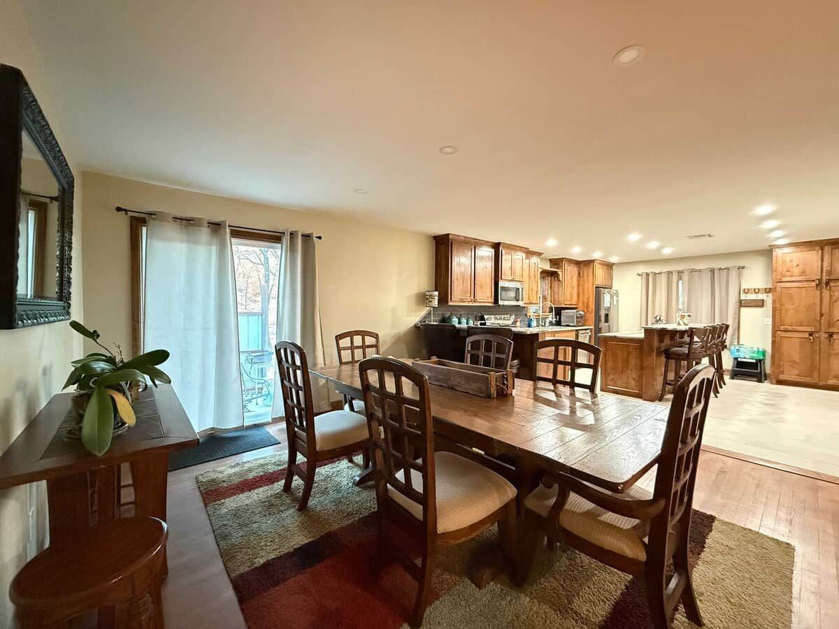 Un comedor con una mesa y sillas de madera, una cocina al fondo y un ventanal corredero que da a un balcón.