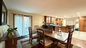 Un comedor con una mesa y sillas de madera, una cocina al fondo y un ventanal corredero que da a un balcón.