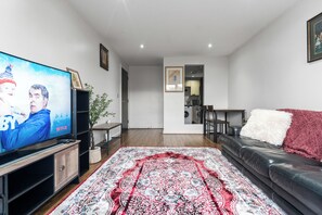 Ein Wohnzimmer mit einem Flachbildfernseher, einem gemusterten Teppich, einer Ledersofa und einem Essbereich im Hintergrund.