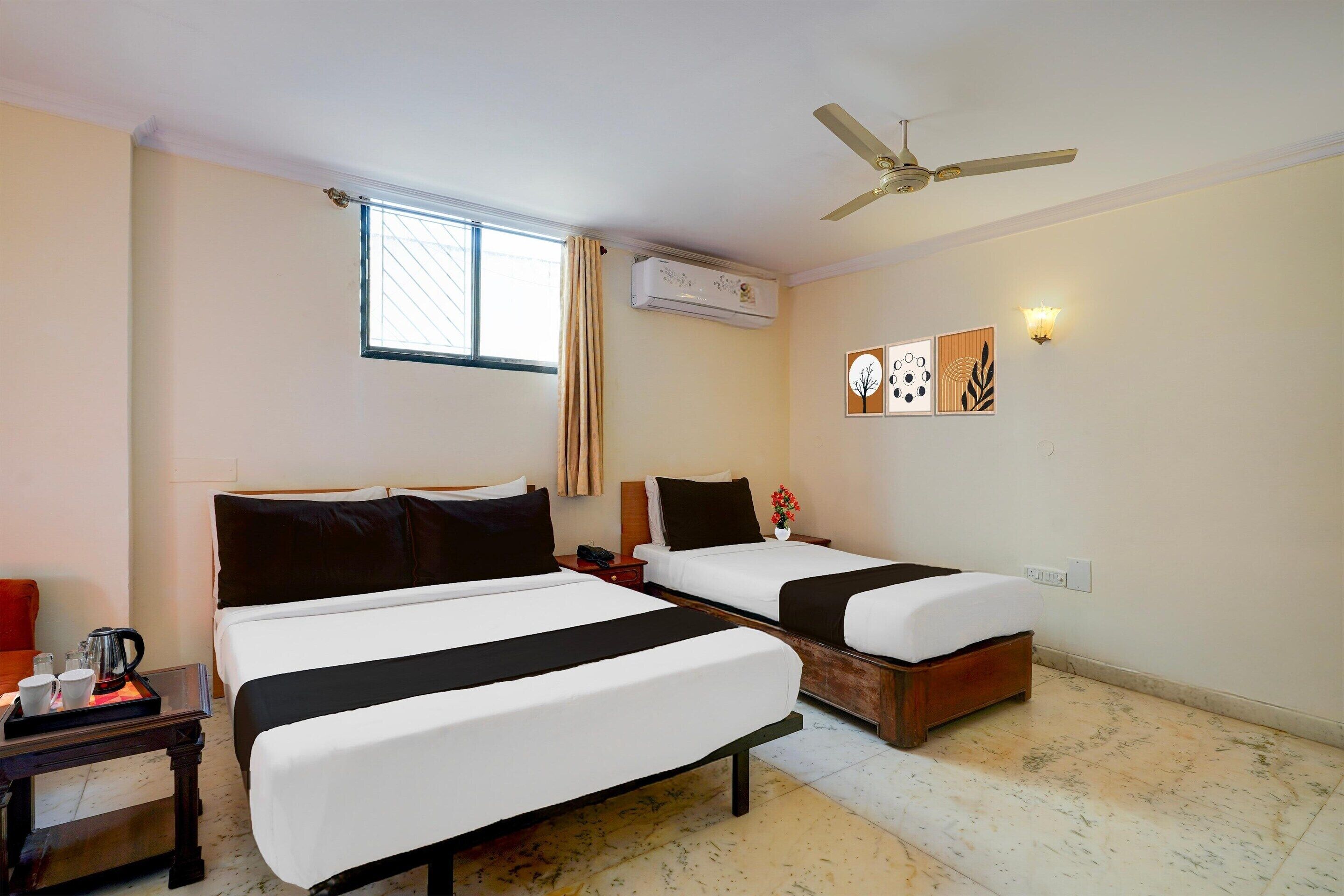 A hotel room with two beds, a ceiling fan, an air conditioning unit, a window with curtains, and a wall-mounted light fixture.