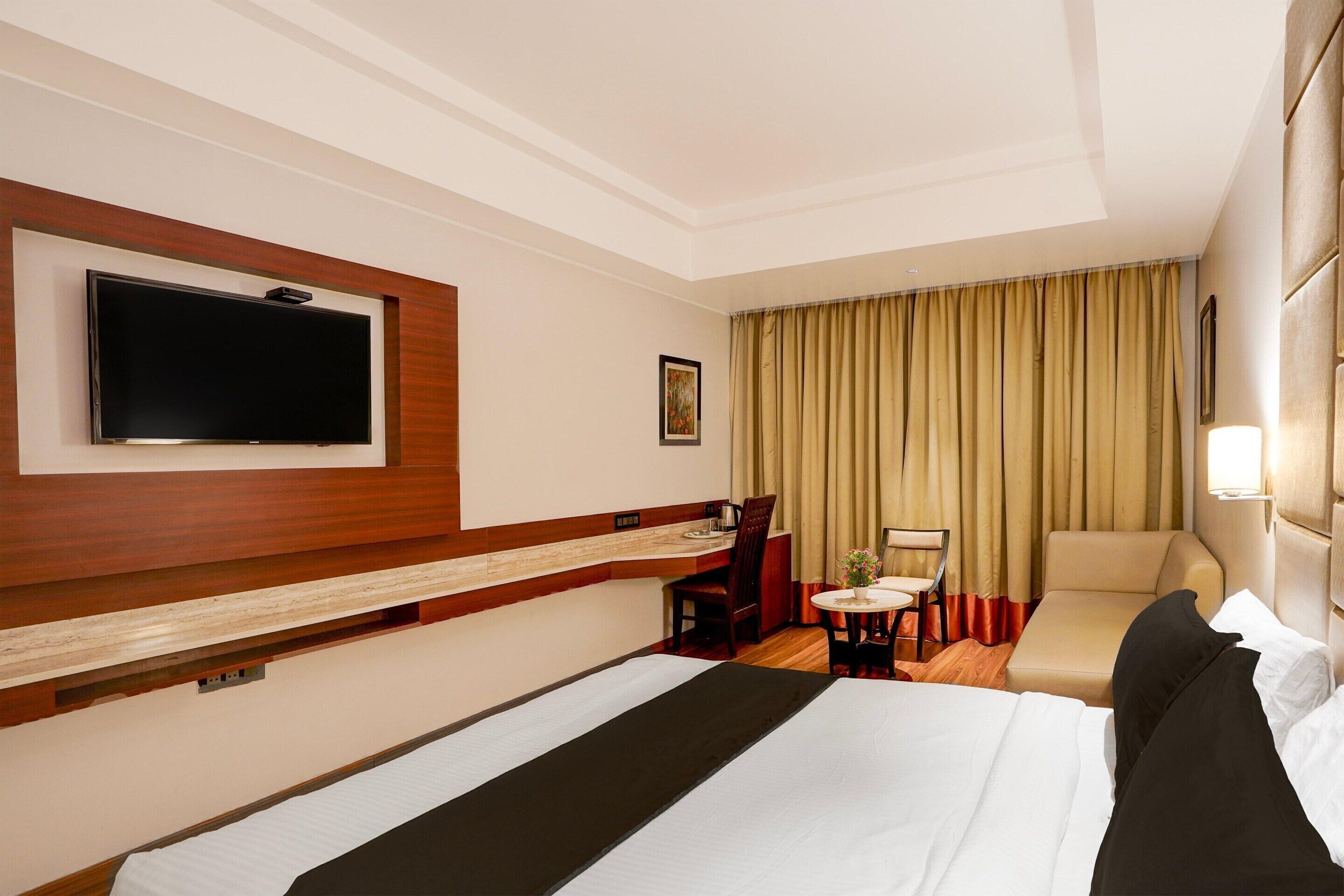 A hotel room with a bed, a TV mounted on the wall, a desk with a chair, a small table with a chair, and a sofa.