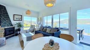 A spacious living room with a fireplace, a sofa, a dining table, and large windows offering a view of the sea.