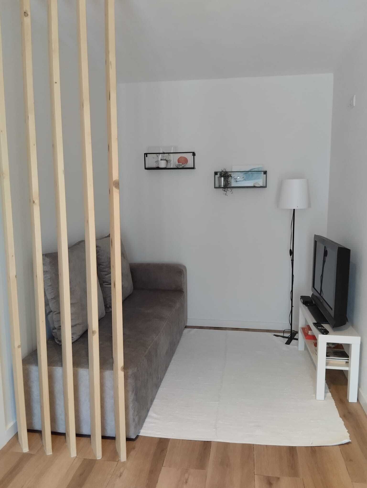 Ein Zimmer mit einem grauen Sofa, einem weißen Teppich, einem Fernseher auf einem weißen Schrank und zwei Wandregalen mit Dekorationselementen.