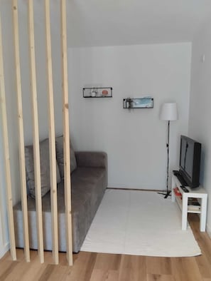 Une pièce avec un canapé gris, un tapis blanc, un téléviseur posé sur un meuble blanc et deux étagères murales ornées d’objets décoratifs.