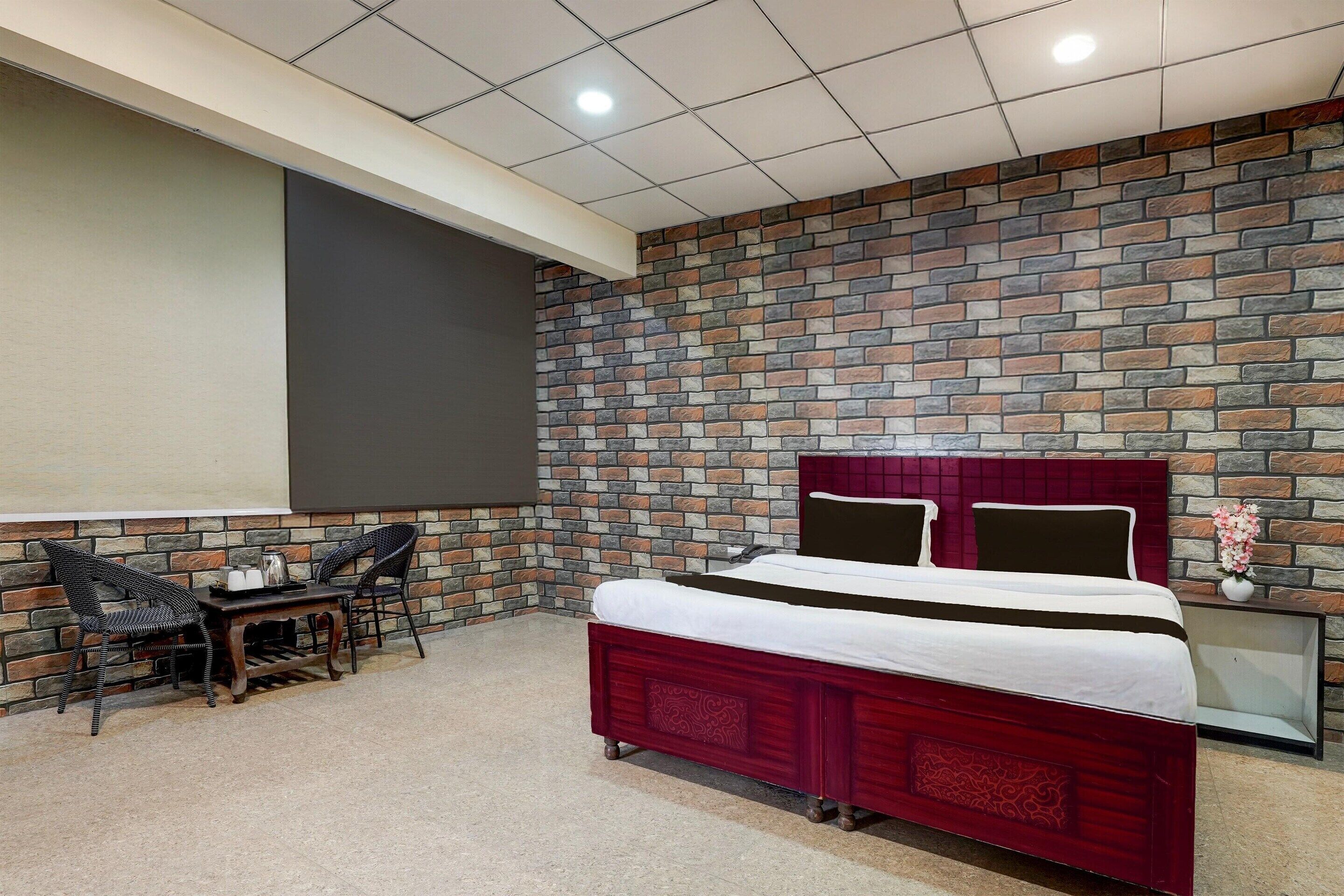 A bedroom with a brick-patterned wall, a bed with a red headboard, two chairs, a small table, and a nightstand with a vase.