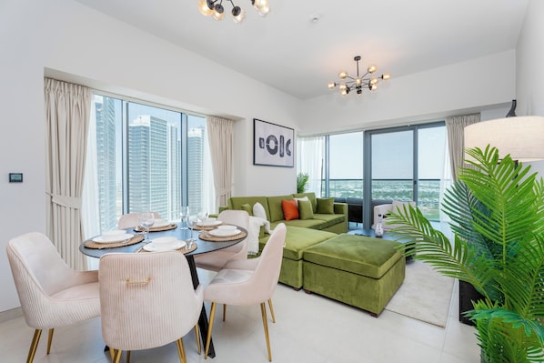 A modern living room with a green sofa, a dining table with chairs, and a large window offering a city view.