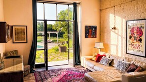 A living room with a sofa, a coffee table, a lamp, and a large window with a view of a garden. - Luxury private condo for 2 w/pool in Apulian hills. Only 15 mins from beach! (Puglia)