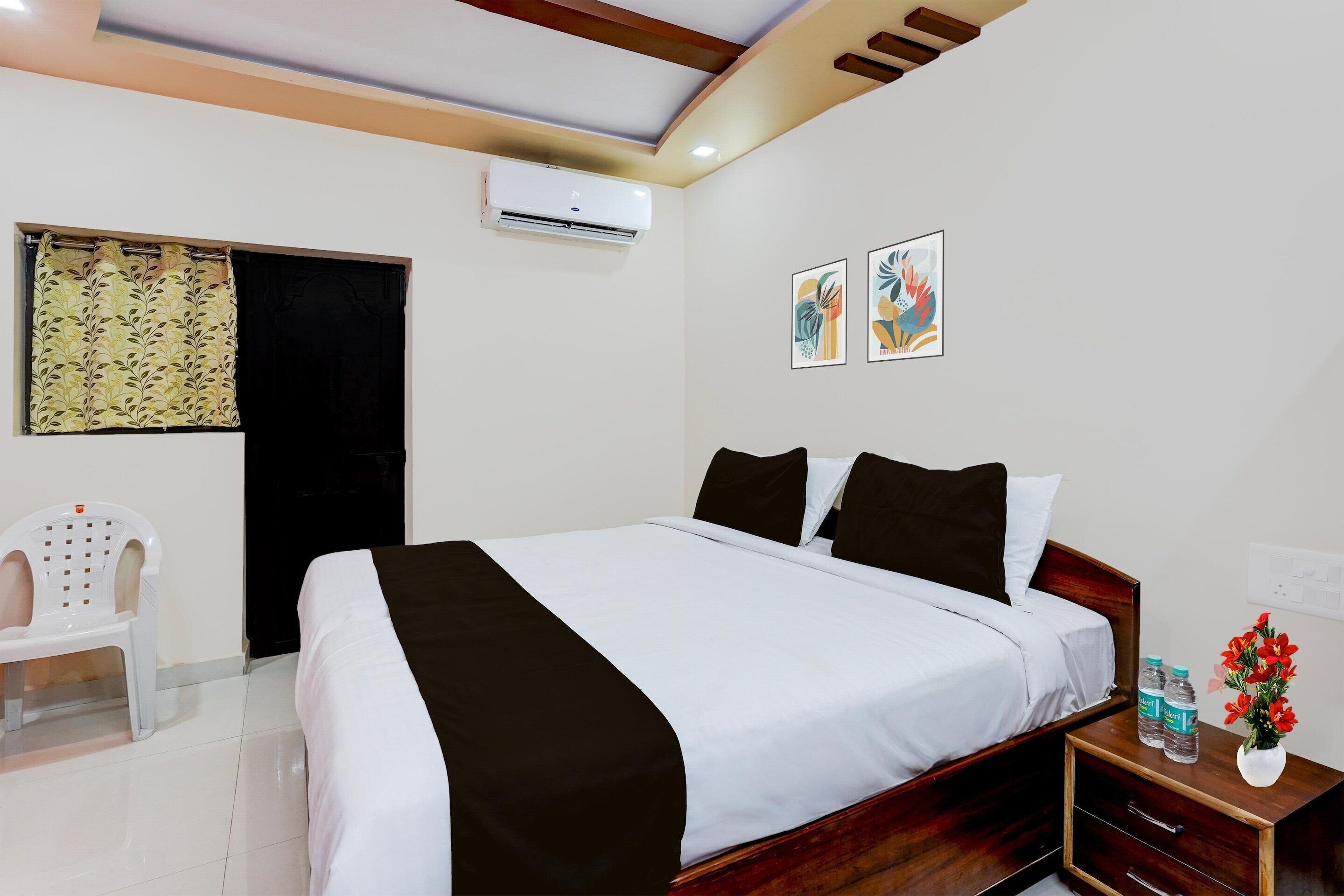 A hotel room with a bed, a nightstand, a chair, an air conditioning unit, and artwork on the wall.