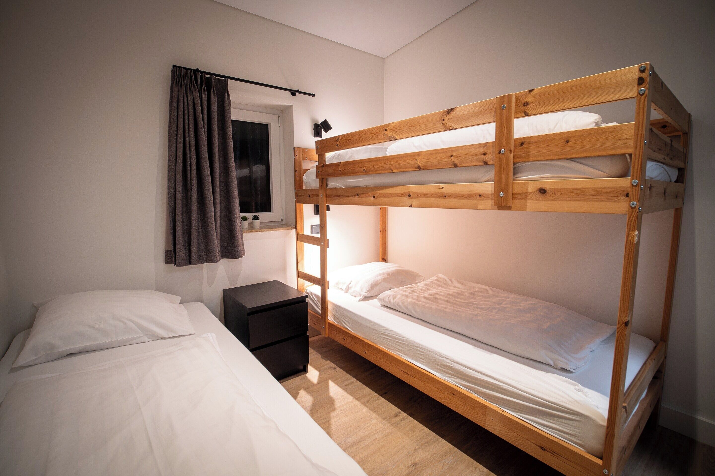 A bunk bed with a wooden frame, a bedside table, a window with curtains, and a white bedspread.