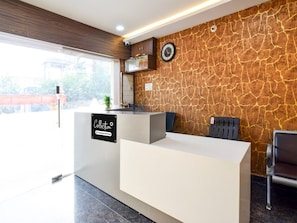 A reception desk with a sign indicating it's a company-serviced collection point, a clock on the wall, and a window with a view of the outside. - Deluxe Hotel Godavari (Hyderabad)