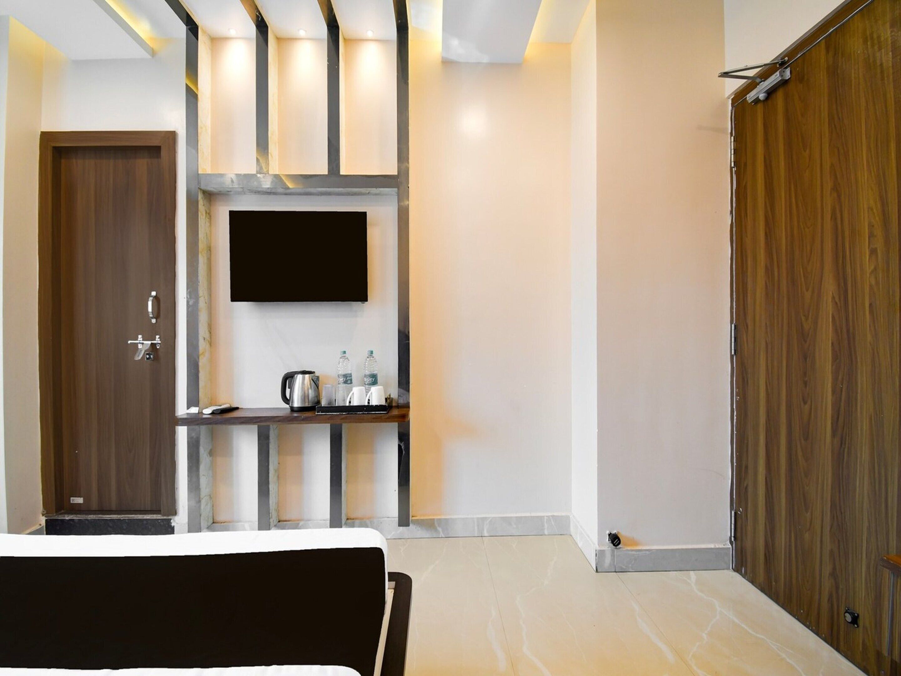 A hotel room with a bed, a TV mounted on the wall, a small table with a kettle and cups, and a wooden door.