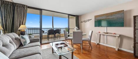 Ein modernes Wohnzimmer mit einer Couch, einem Glastisch, einem Essbereich mit Stühlen, einem an der Wand befestigten Fernseher und einem Balkon mit Blick.