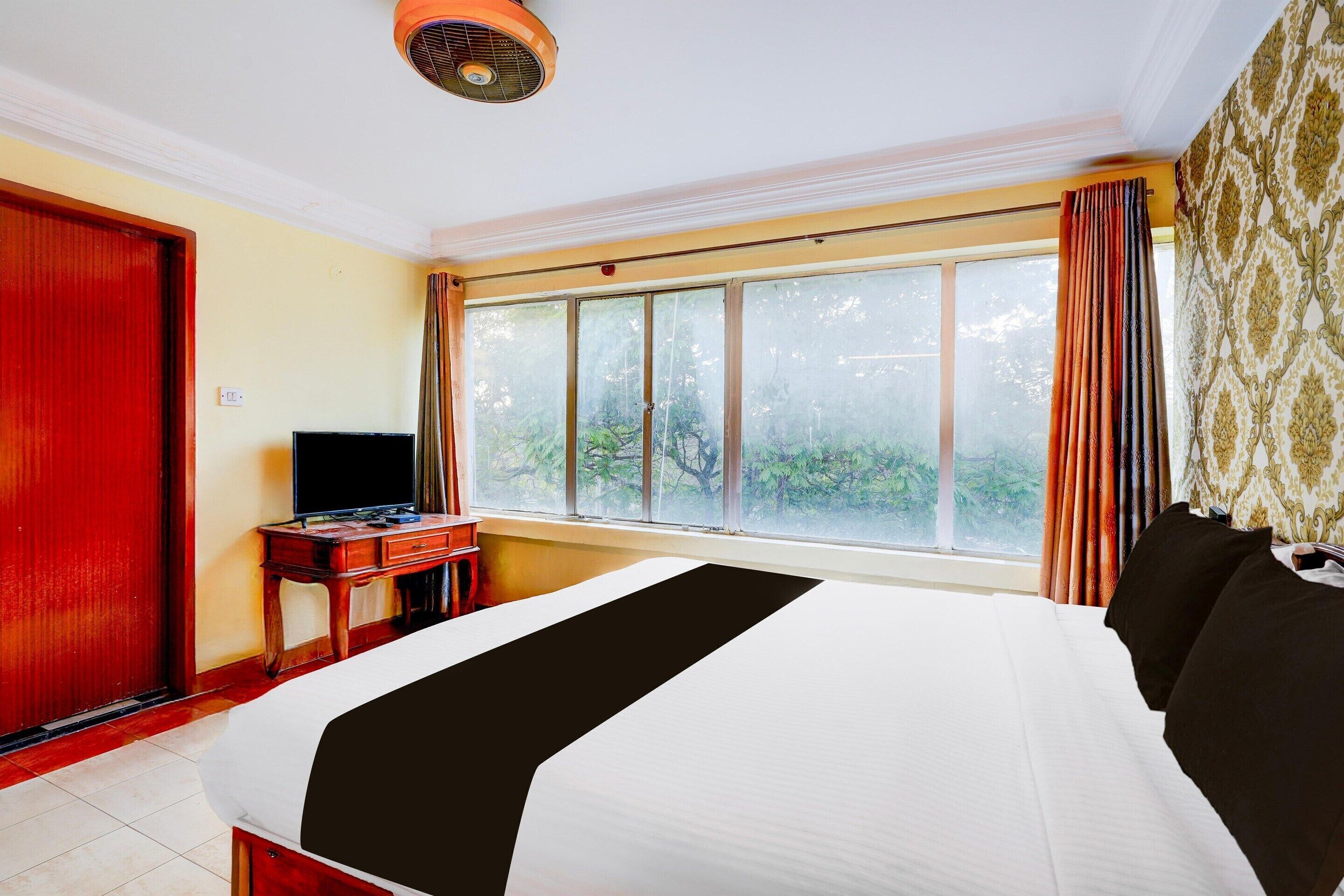 A hotel room with a bed, a desk with a television, a window with curtains, and a ceiling fan.