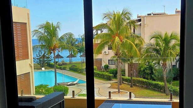 Una habitación con un amplio ventanal corredero que da a una zona de piscina con palmeras y vistas al mar.