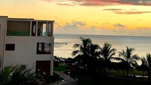 Un edificio moderno con varios balcones que dan a una masa de agua, palmeras en primer plano y un cielo al atardecer.