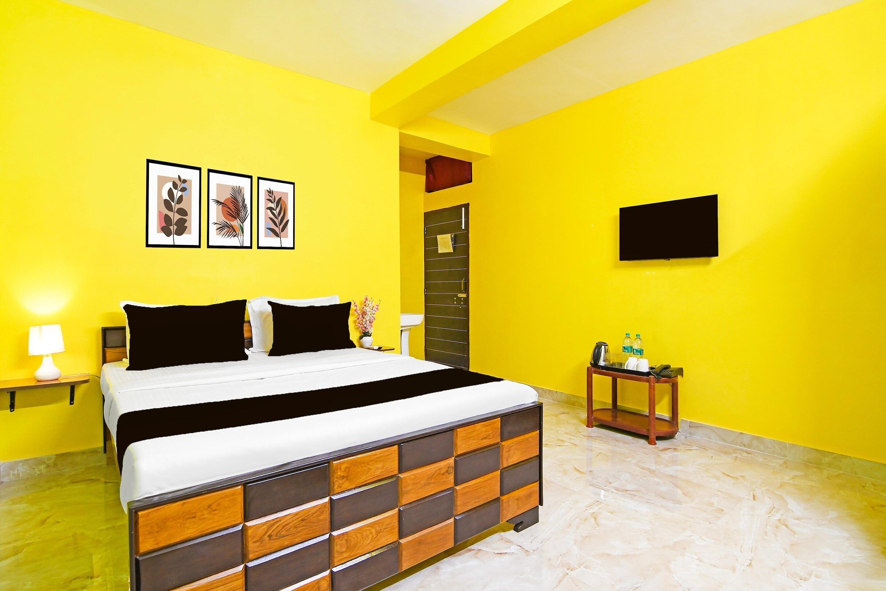 A hotel room with a bed, a bedside table, a small table with bottles, a TV, and framed artwork on the wall.