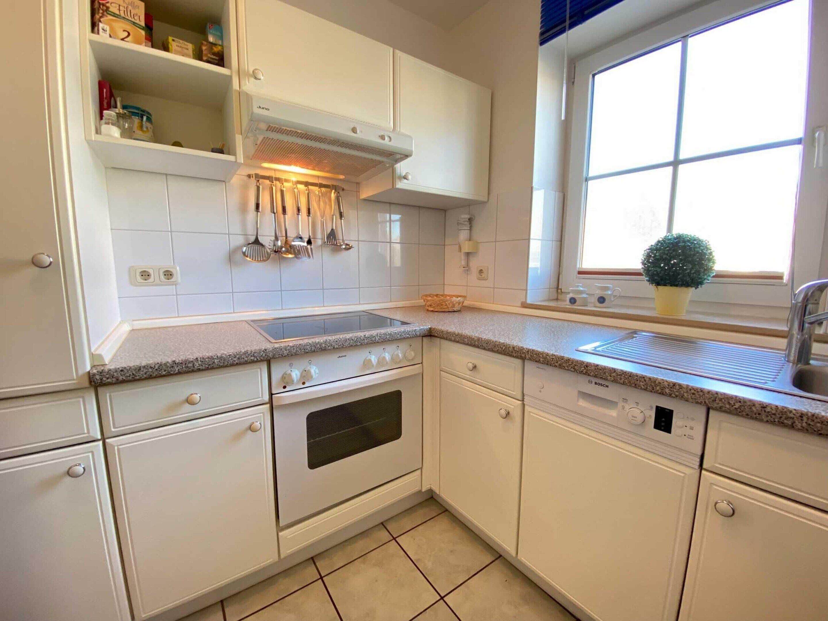 A kitchen with white cabinets, a stove, oven, and dishwasher. There is a window with a plant on the windowsill.