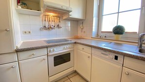 Une cuisine avec des meubles blancs, une cuisinière, un four et un lave-vaisselle. Il y a une fenêtre avec une plante posée sur le rebord.